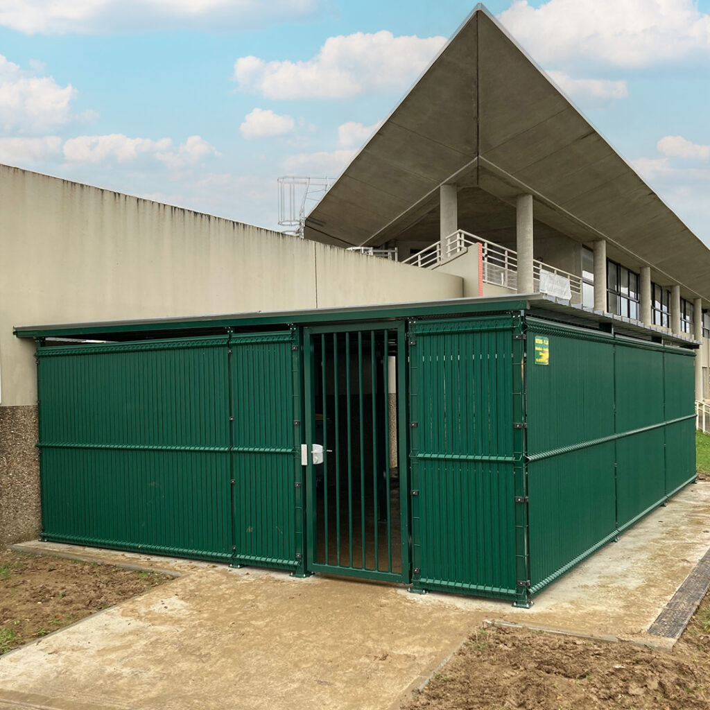 Abri sécurisé en acier thermolaqué vert, avec porte grillagée, installé sur dalle béton devant un bâtiment tertiaire moderne.
