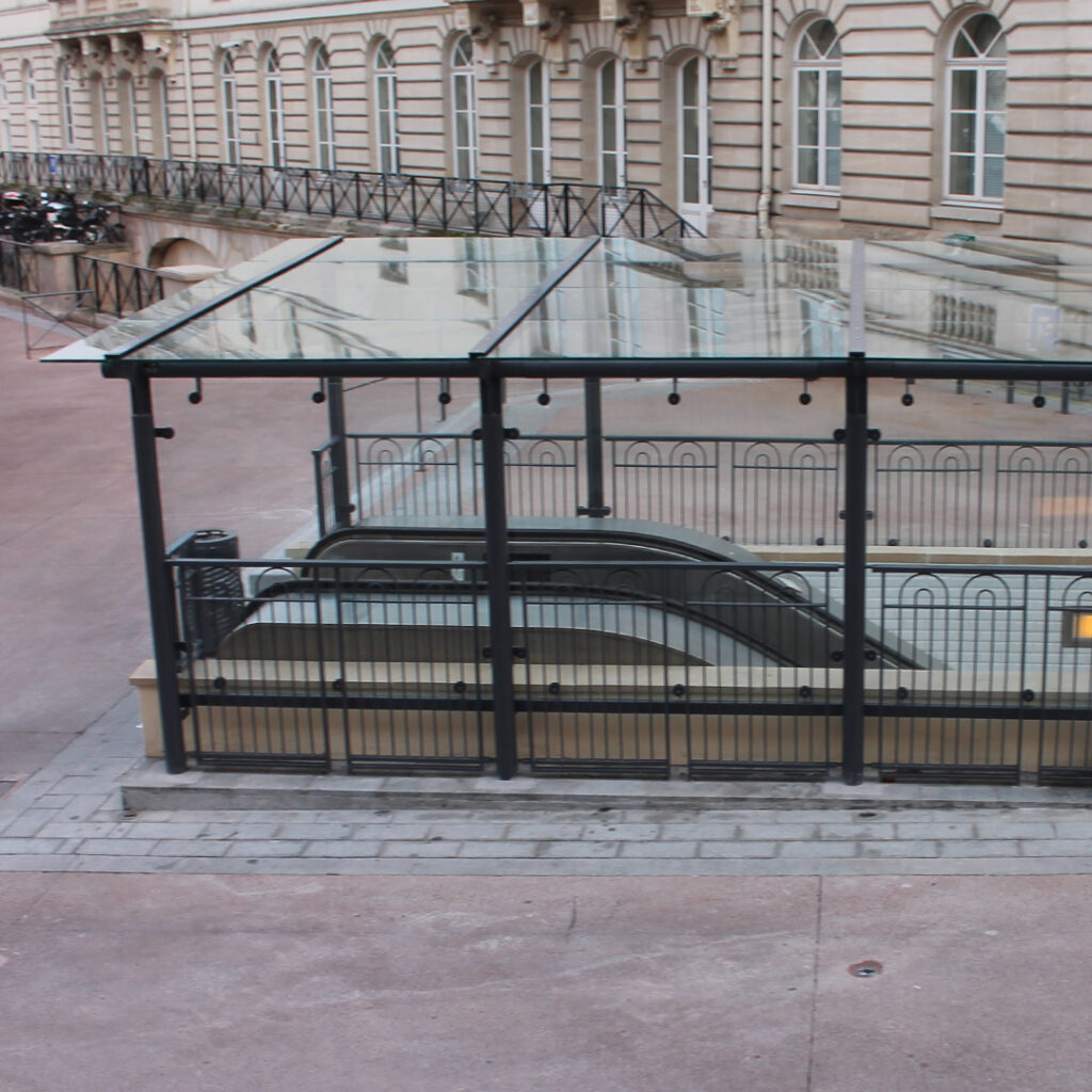 Abri en verre et acier protégeant une sortie de métro équipée d’escalators.