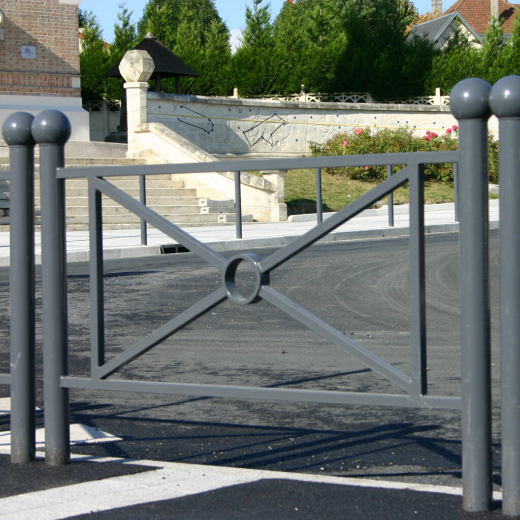 Barrière urbaine en acier thermolaqué gris, avec motif décoratif central, installée pour sécuriser et structurer un espace public paysager.