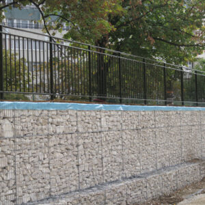 Mur de soutènement en gabion surmonté d’une clôture barreaudée noire en acier.