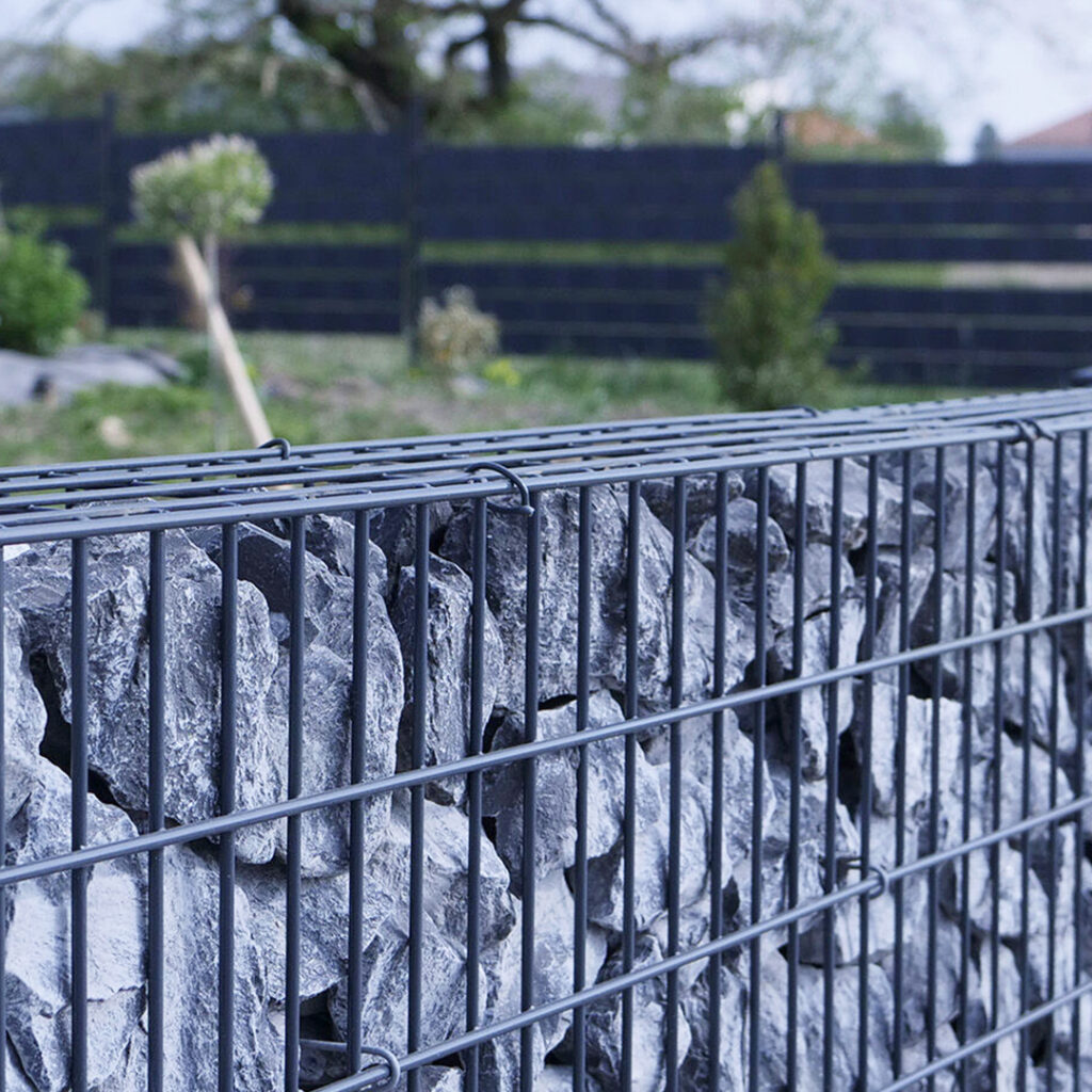 Clôture en gabion remplie de pierres grises, installée dans un jardin paysager.