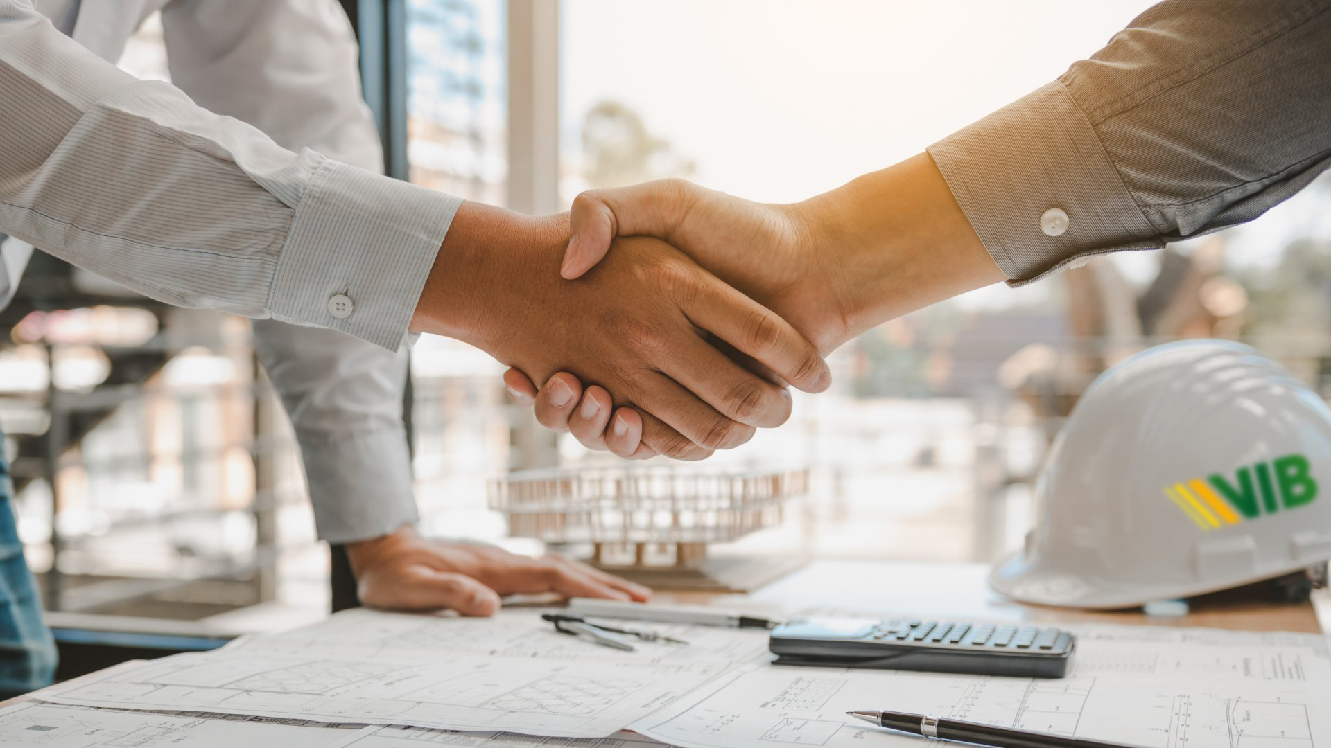 Poignée de main entre deux personnes sur un bureau avec des plans techniques et un casque de chantier VIB.