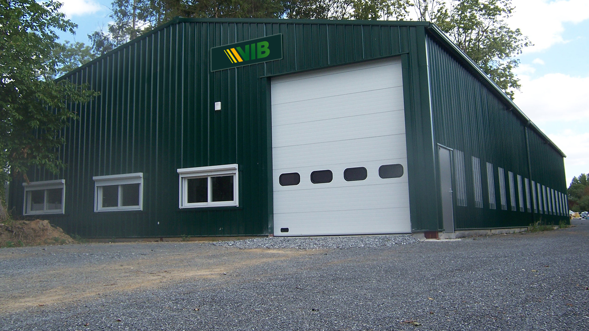 Façade du bâtiment de l'entreprise de métallerie VIB, avec bardage métallique vert, grande porte sectionnelle blanche et accès latéral sur site arboré.