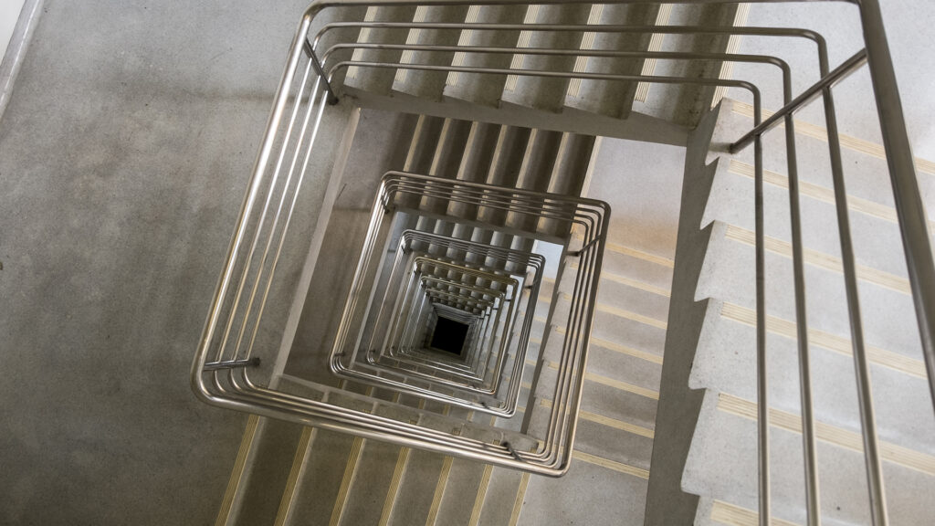 Vue plongeante d’un escalier intérieur en béton avec mains courantes continues en inox, fixées sur les murs d’une cage d’escalier.
