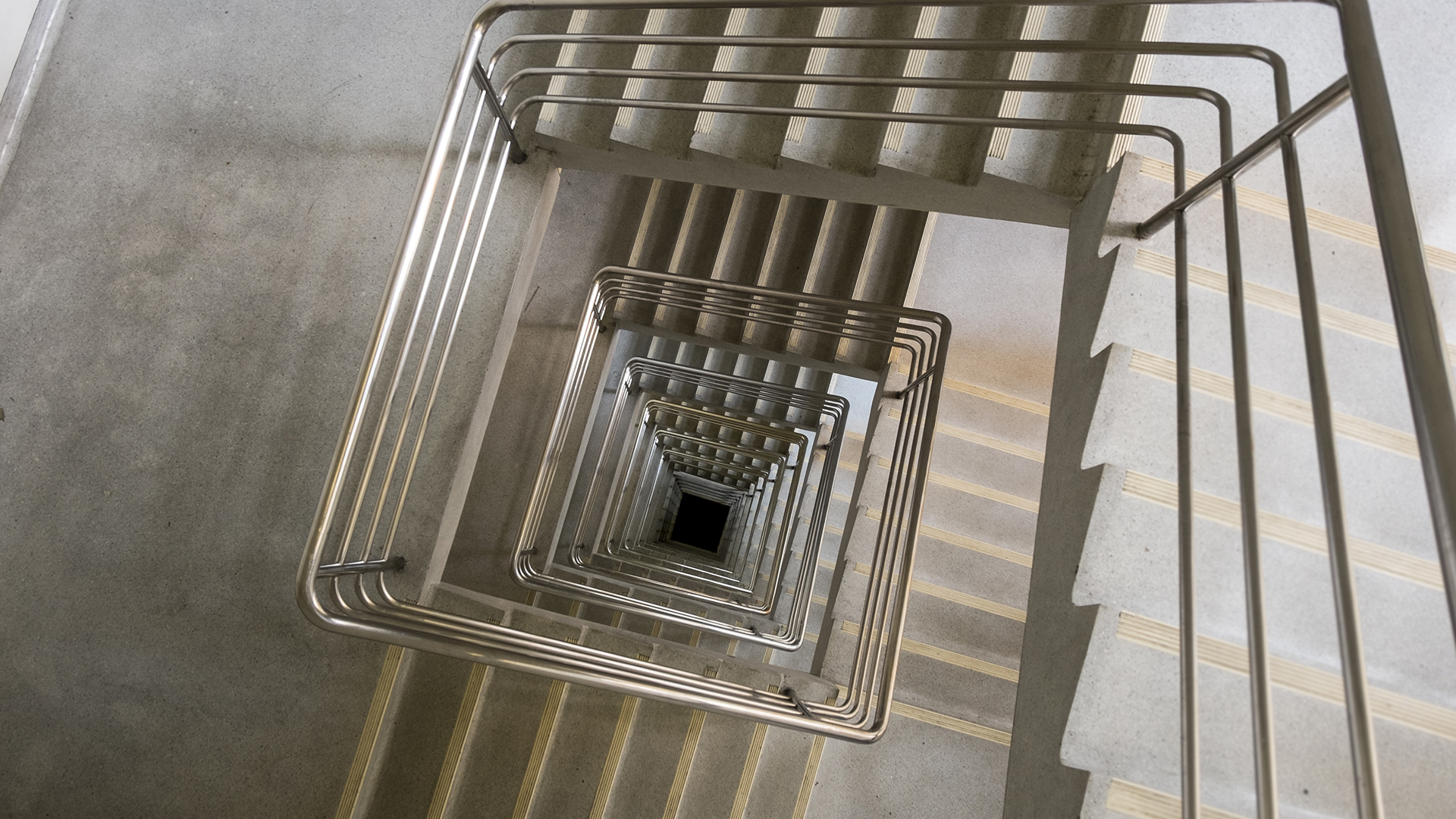 Vue plongeante d’un escalier intérieur en béton avec mains courantes continues en inox, fixées sur les murs d’une cage d’escalier.