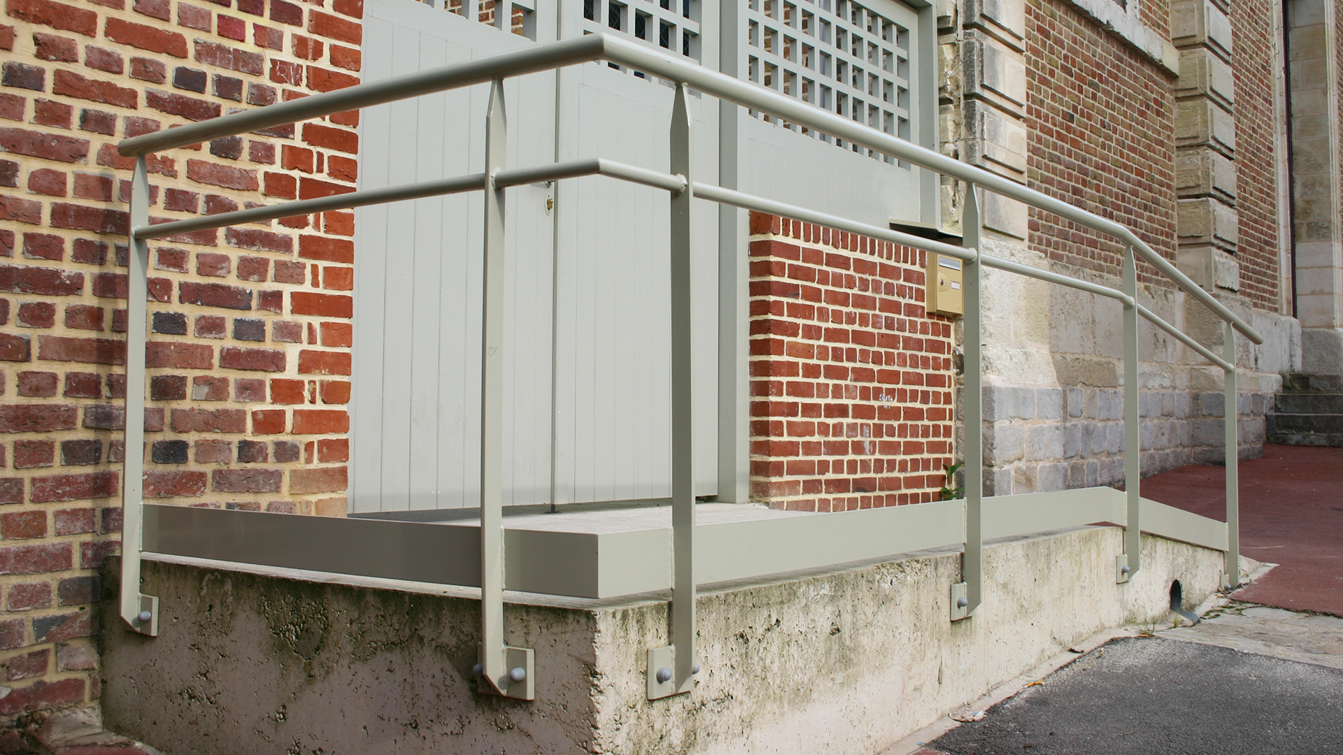 Rampe d’accès PMR avec garde-corps en acier thermolaqué, fixée sur muret béton, installée en façade d’un bâtiment en briques et pierre.