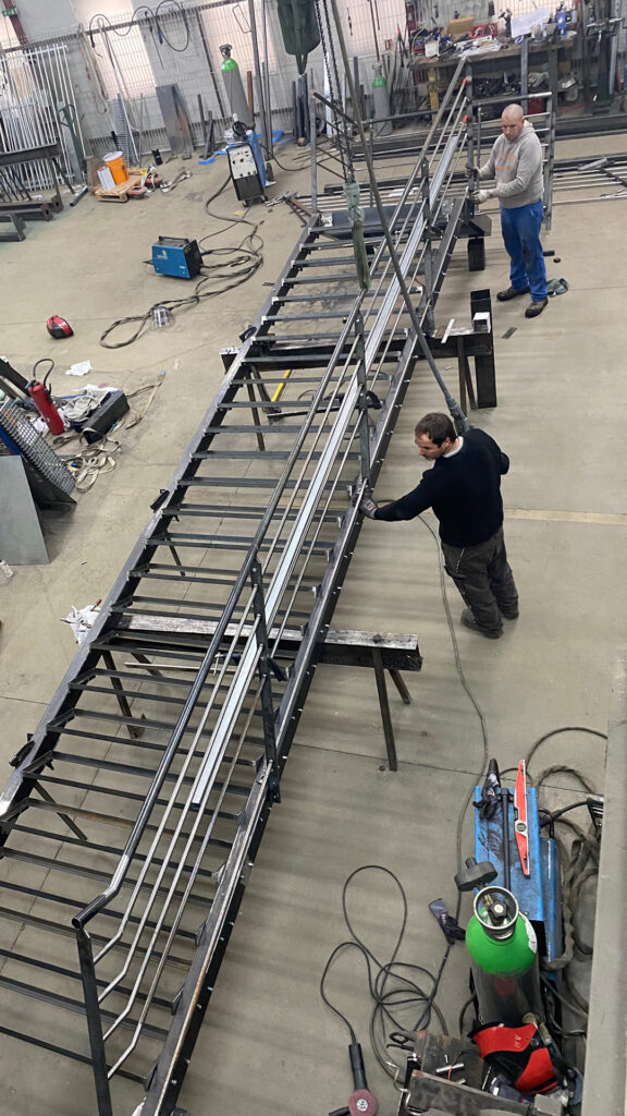 Assemblage d’un escalier métallique avec garde-corps dans un atelier, en cours de finition par deux opérateurs à l’aide d’outils de soudure et de ponçage.
