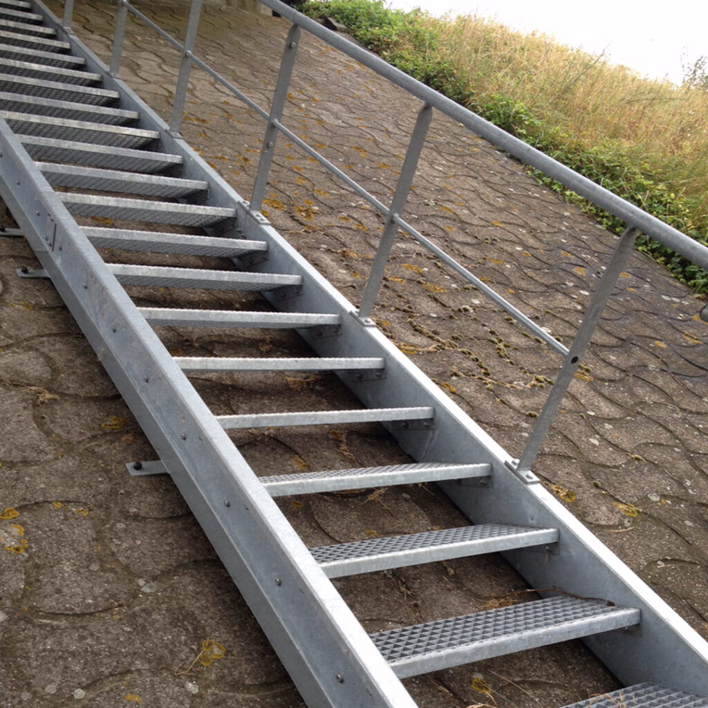 Escalier métallique galvanisé posé sur talus en pente, avec marches ajourées et main courante.