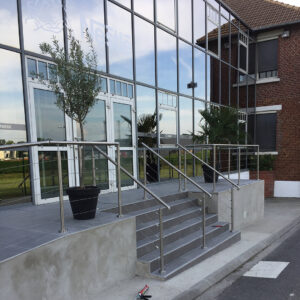 Entrée de bâtiment avec escalier en béton et garde-corps inox poli.