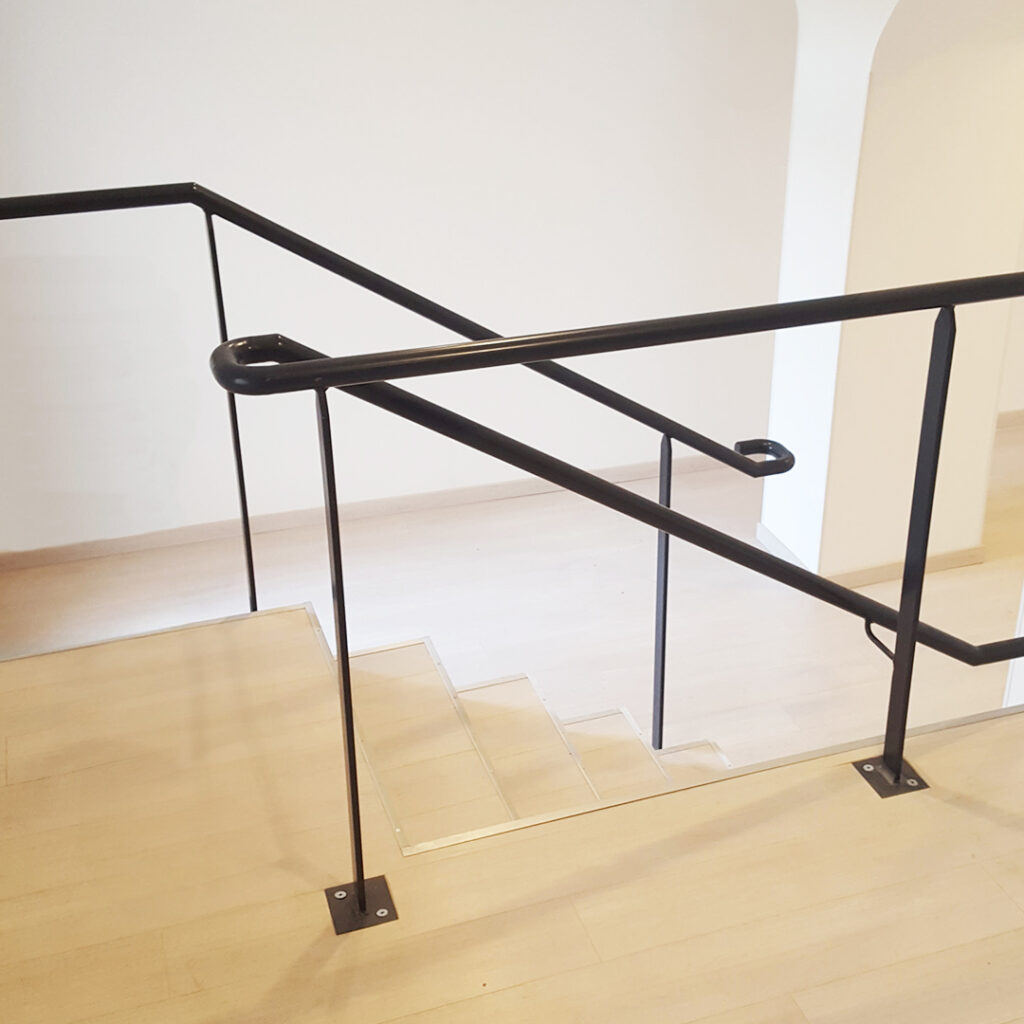 Main courante en acier thermolaqué noir, fixée au sol le long d’un escalier intérieur en bois clair, dans un bâtiment public ou tertiaire.