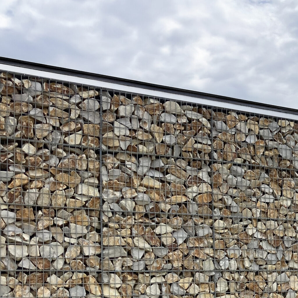 Mur en gabion rempli de pierres naturelles de couleur ocre et grise, sous un ciel nuageux.