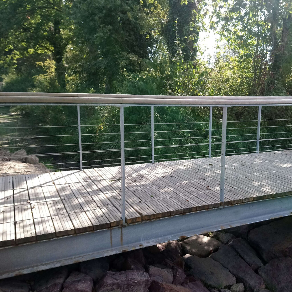 Passerelle piétonne en bois avec garde-corps en câble inox, traversant une zone boisée.