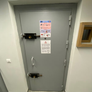 Porte blindée grise avec deux verrous de sécurité, affiches d'instruction et poignée renforcée.