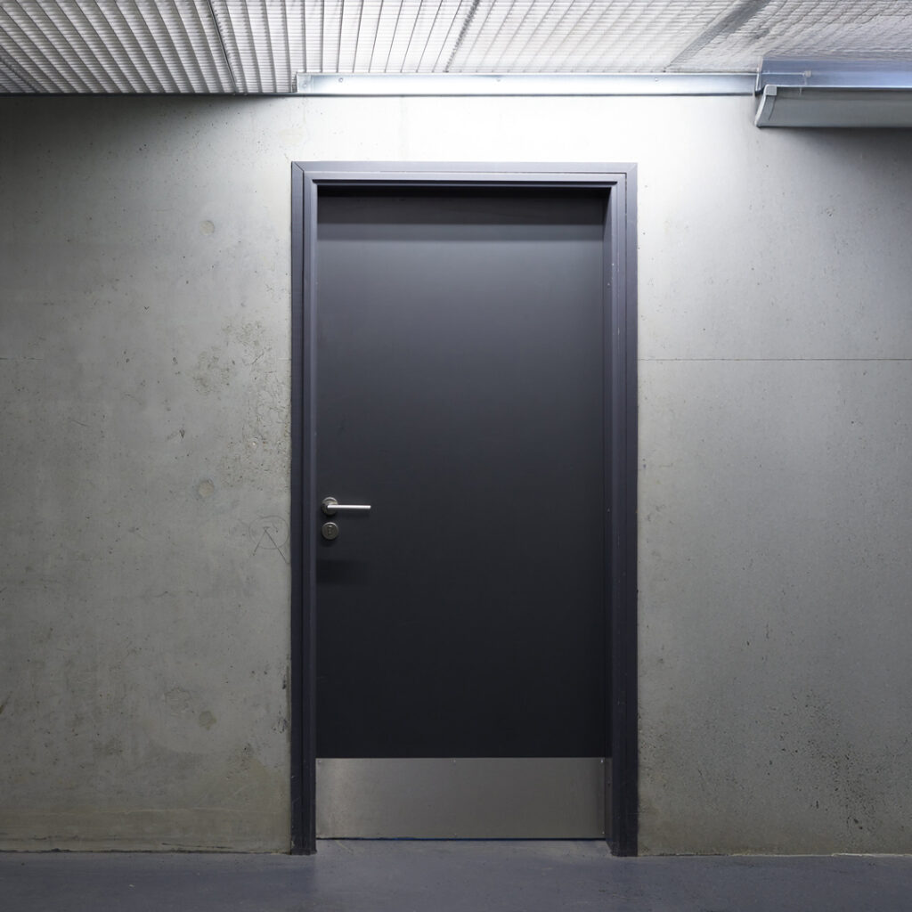 Porte technique pleine en acier, finition gris anthracite, avec plinthe inox en partie basse, installée dans un couloir de service ou local technique.