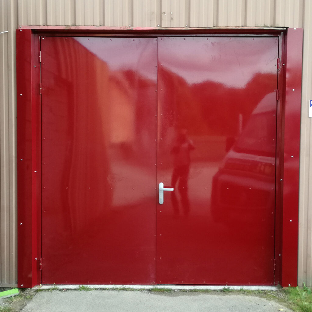 Porte métallique rouge à double battant, avec poignée centrale, installée sur la façade d’un bâtiment à usage industriel ou technique.