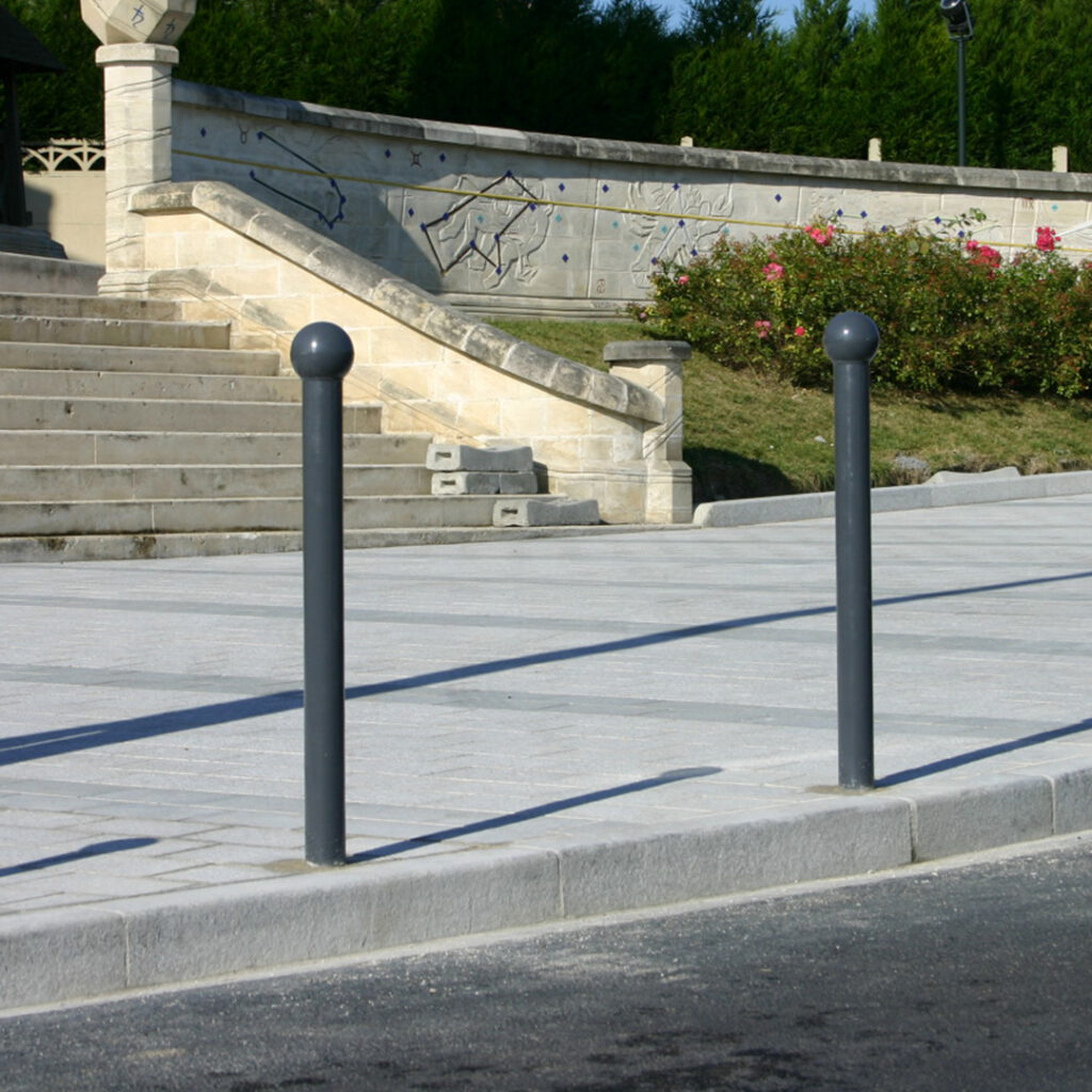 Bornes de voirie en acier installées en bordure de trottoir pour sécuriser une zone piétonne face à un escalier en pierre et un mur ornementé.