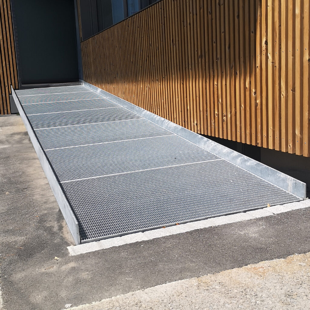 Rampe d’accès PMR en caillebotis acier galvanisé, installée le long d’un bâtiment en bois pour faciliter l’accessibilité aux personnes à mobilité réduite.