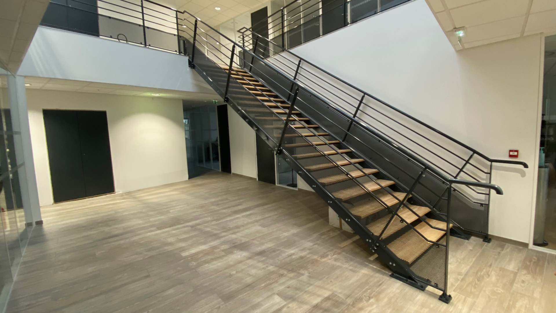Escalier intérieur en métal et bois dans le hall d’un bâtiment moderne, avec garde-corps design noir.