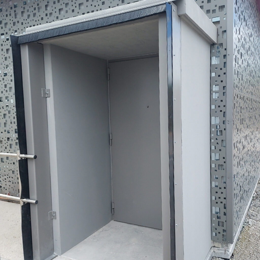 Sas d’accès sécurisé en métal avec porte blindée grise, intégré à une façade de bâtiment à parement perforé.